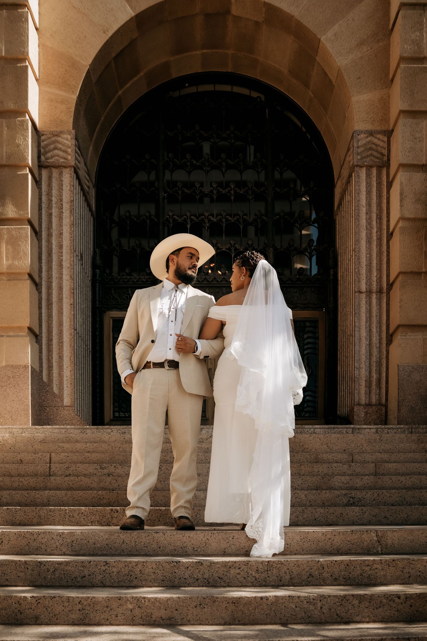 Bride and groom stand on stone steps before arched doorway, she in white gown with veil, he in tan suit and cowboy hat.