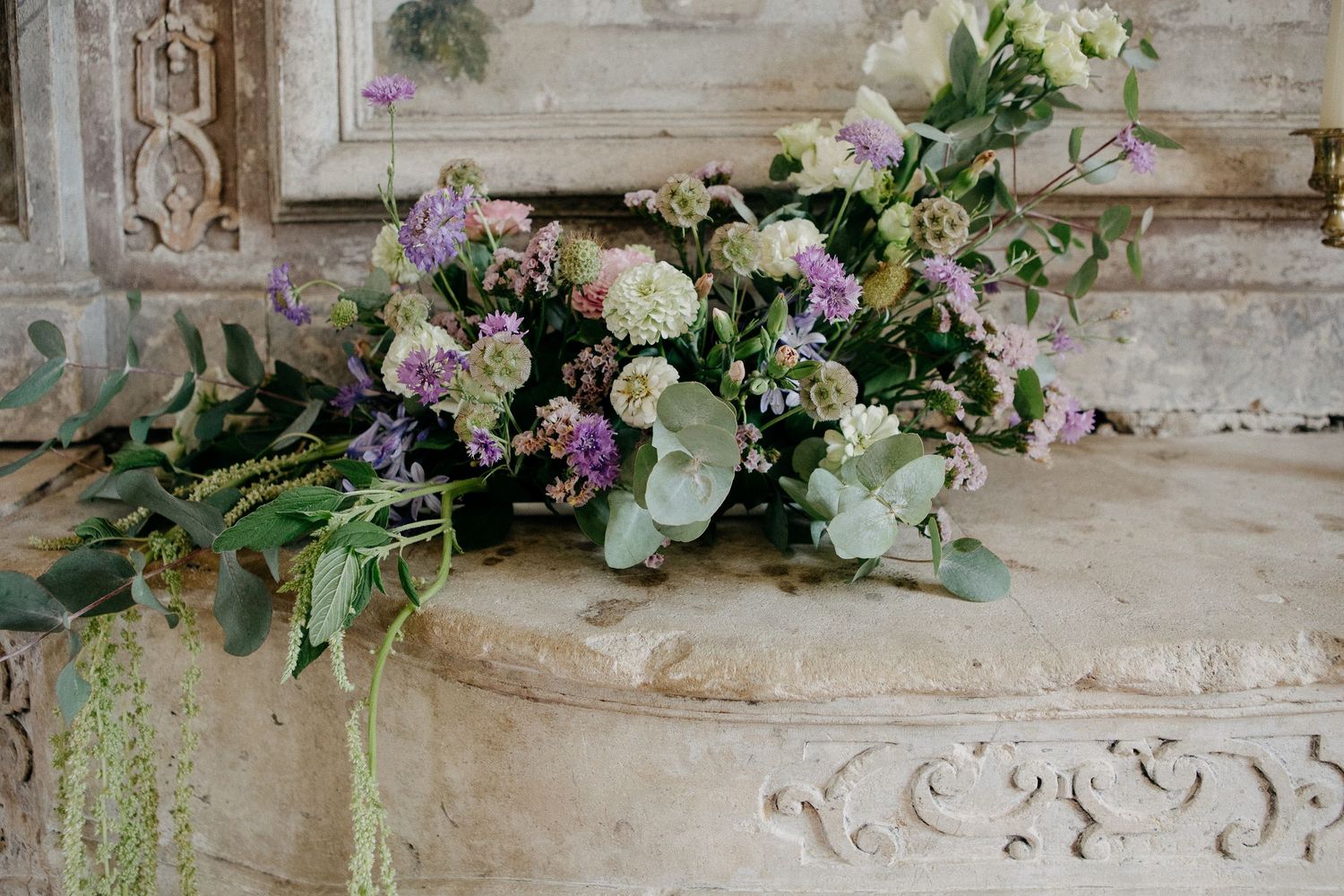 Elegant floral arrangement with purple, white blooms and eucalyptus on ornate stone mantel.