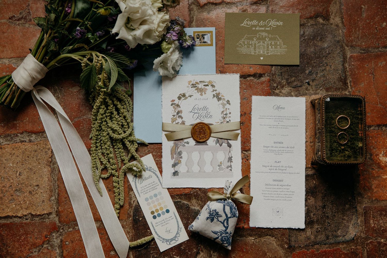 Elegant wedding stationery flat lay with floral invitations, wax seal, menu cards, and bridal bouquet on rustic brick surface.