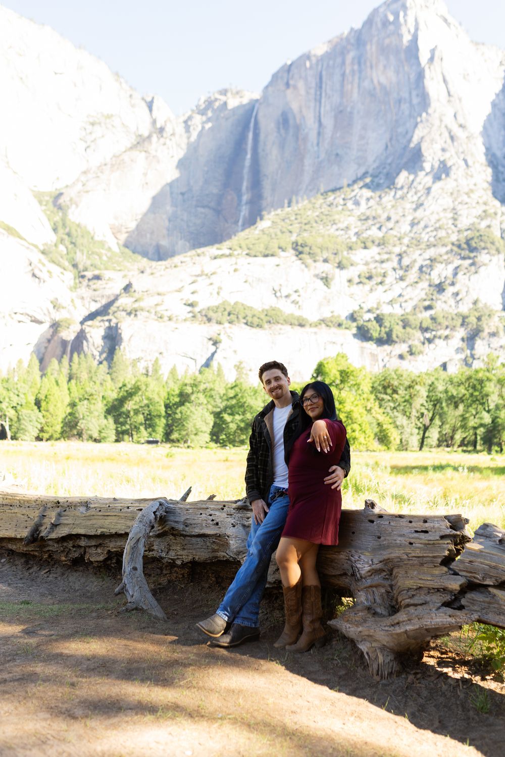 Couple posing on a fallen log with Yosemite Falls and granite cliffs in the background on a sunny day.
