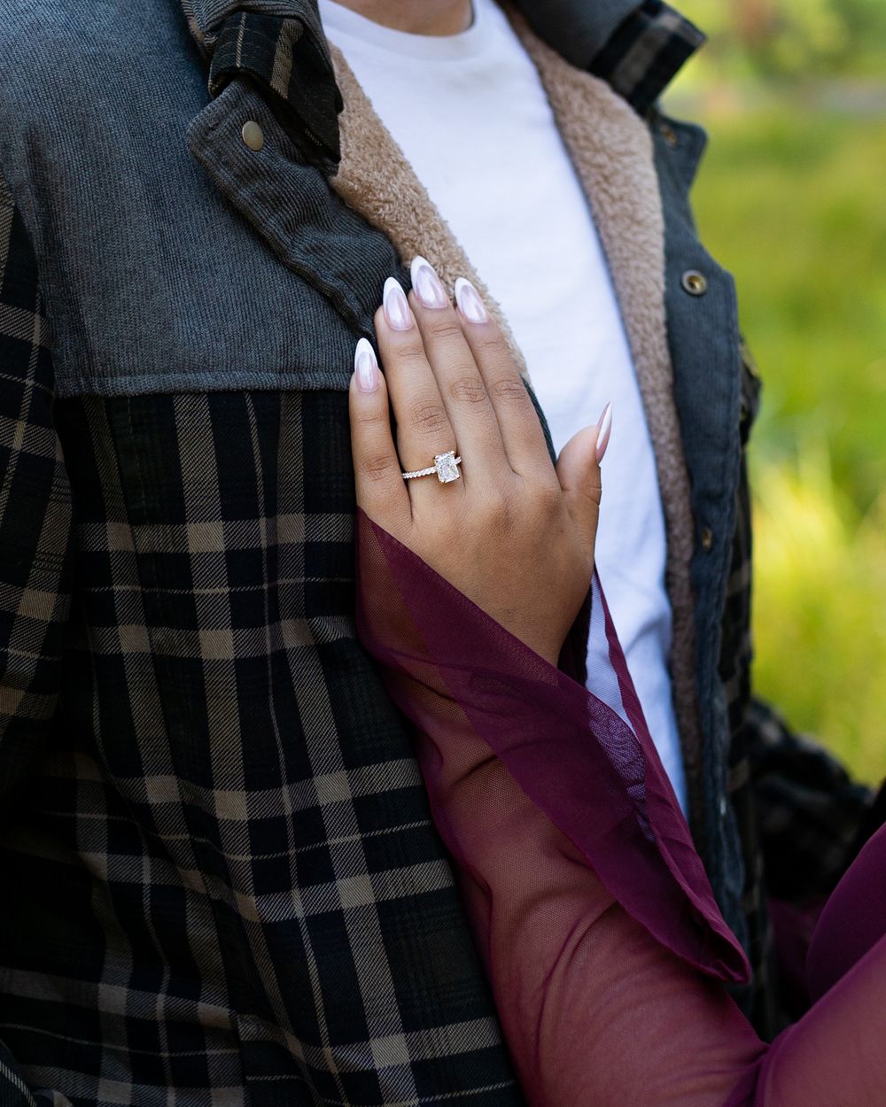 Close-up of a hand with white nails displaying a diamond engagement ring, resting on a plaid jacket outdoors.