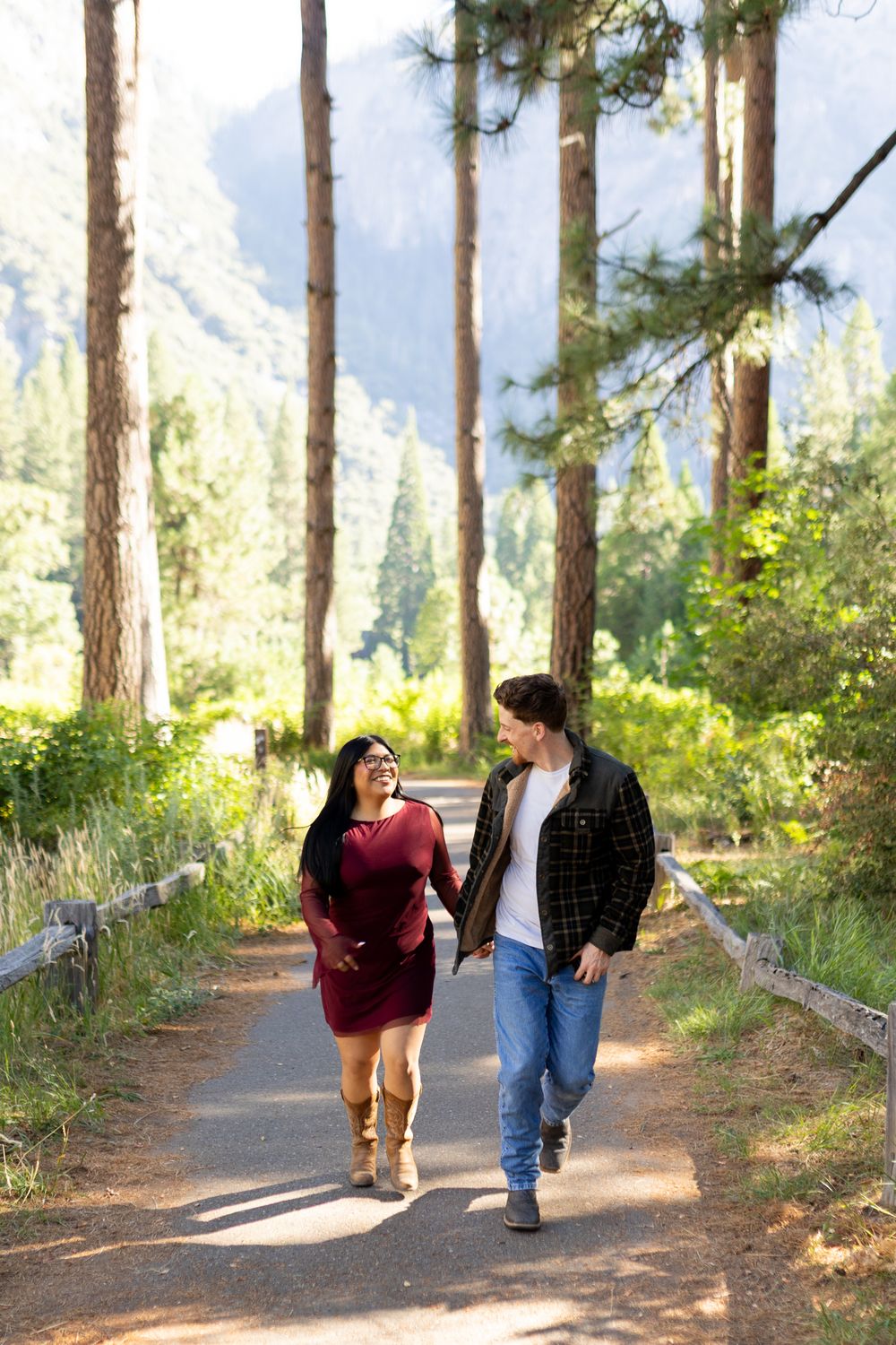 Couple walking hand-in-hand on a forest path surrounded by tall pine trees and mountains in the background.