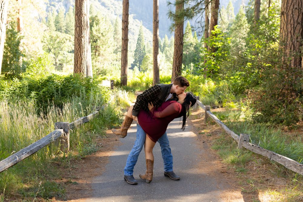 Couple sharing a romantic dip kiss on a forest path surrounded by tall pine trees and lush greenery.