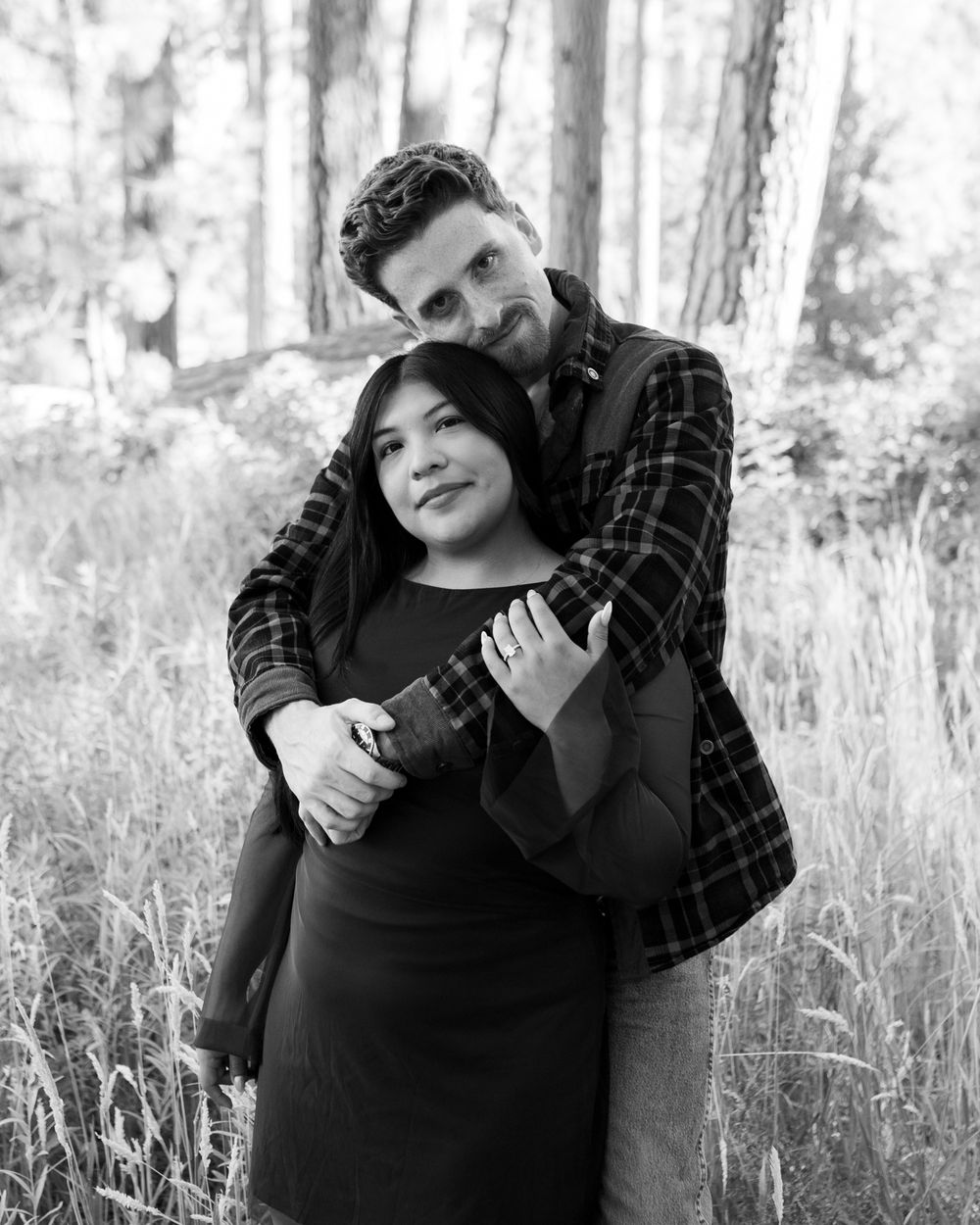 Black and white photo of a couple embracing in a forest, woman wearing a ring, man hugging her from behind.