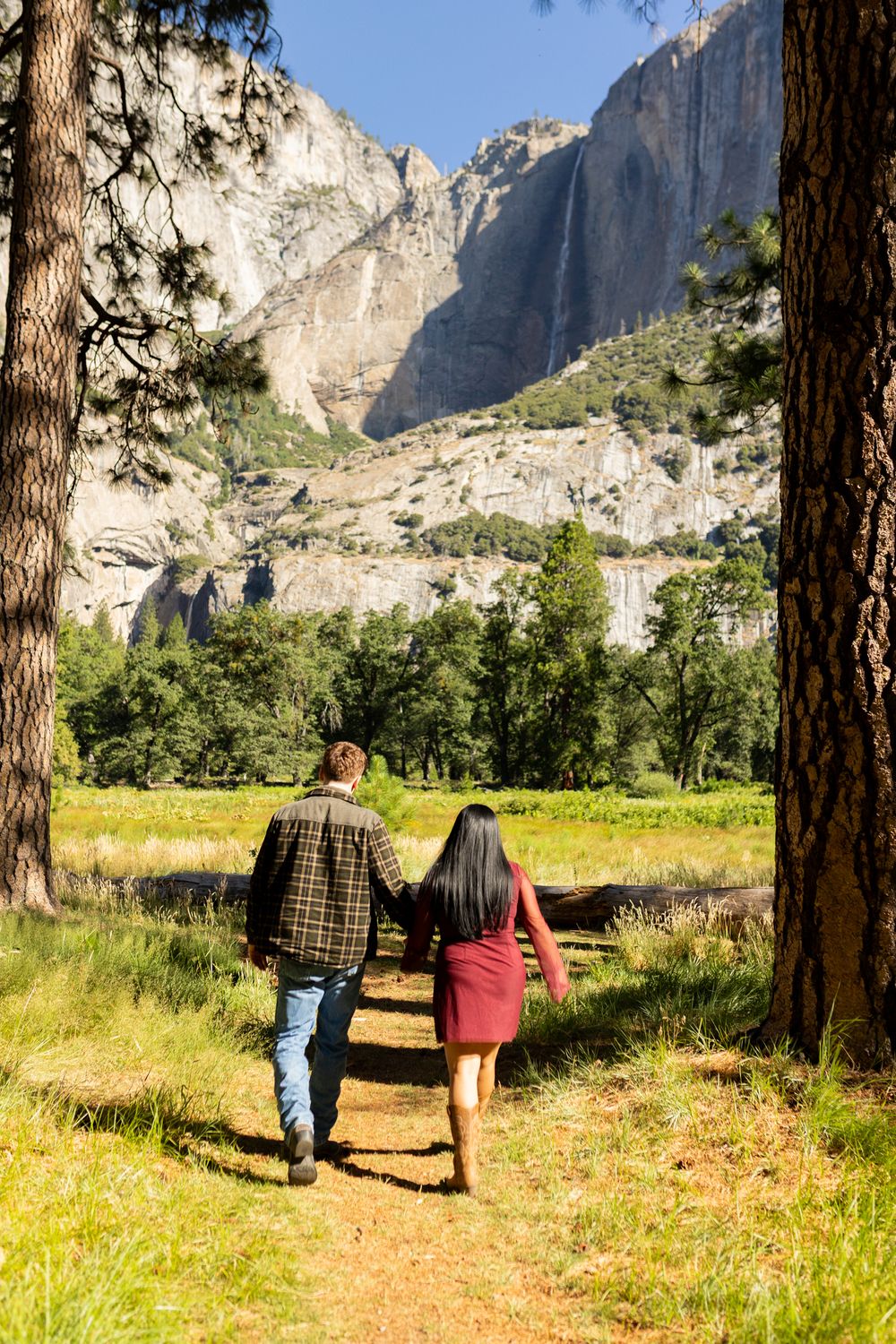 Couple holding hands walking on a trail in Yosemite Valley with granite cliffs and waterfall in background.