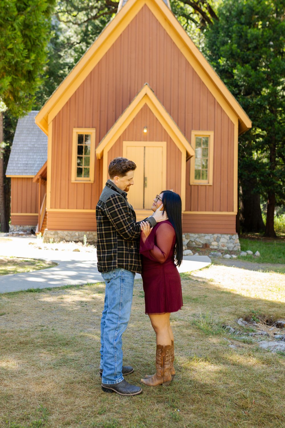 Couple holding hands facing each other in front of a rustic brown yosemite park chapel outdoors.