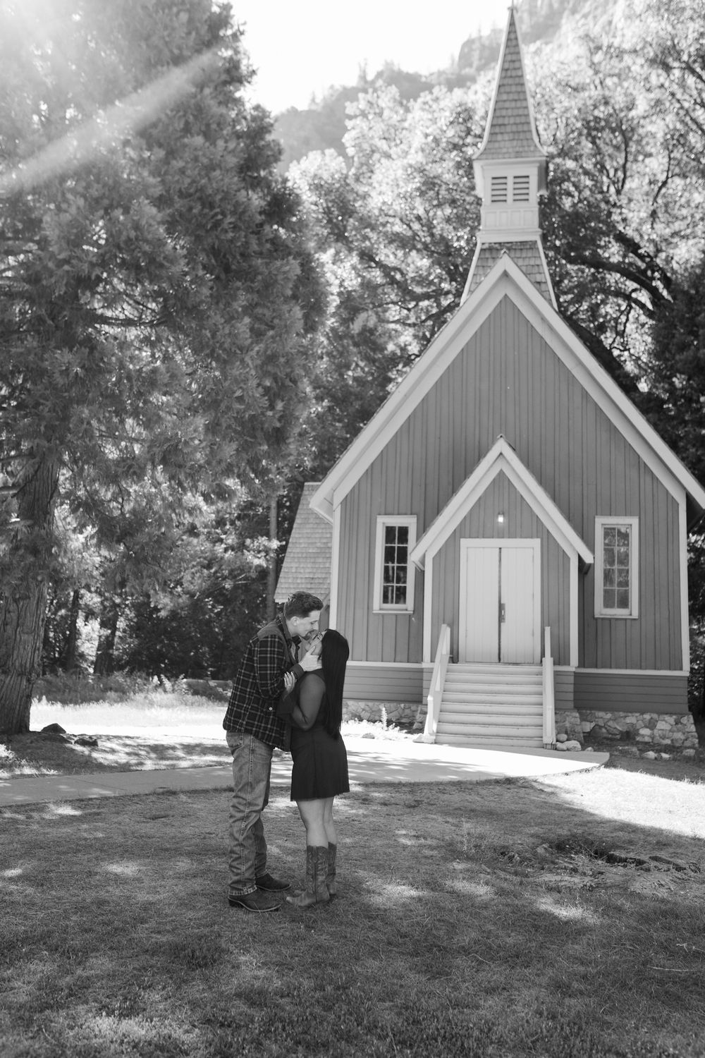 Couple embracing in front of a charming wooden chapel surrounded by trees in a black and white photo.