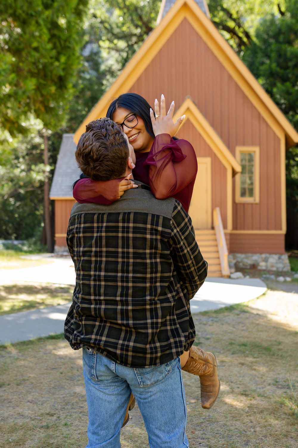 A couple embraces outdoors near a rustic wooden chapel, as the woman shows off her engagement ring.