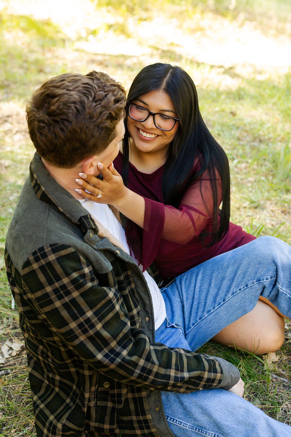 A smiling young woman with glasses touches a young man's face as they sit together outdoors on grass.