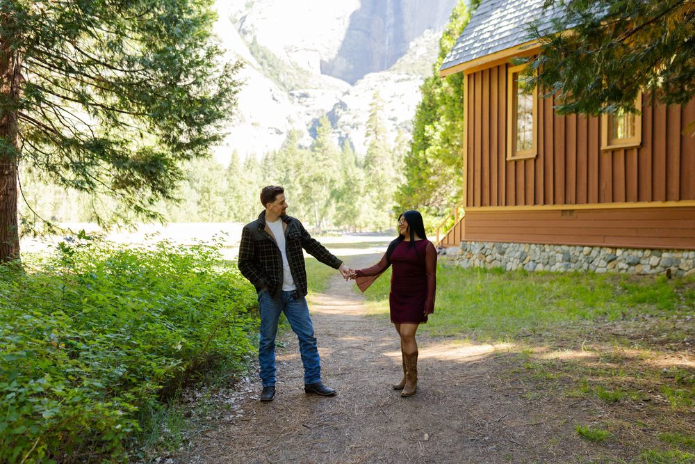 Couple holding hands on a forest path beside a rustic cabin with mountain views in a scenic outdoor setting.