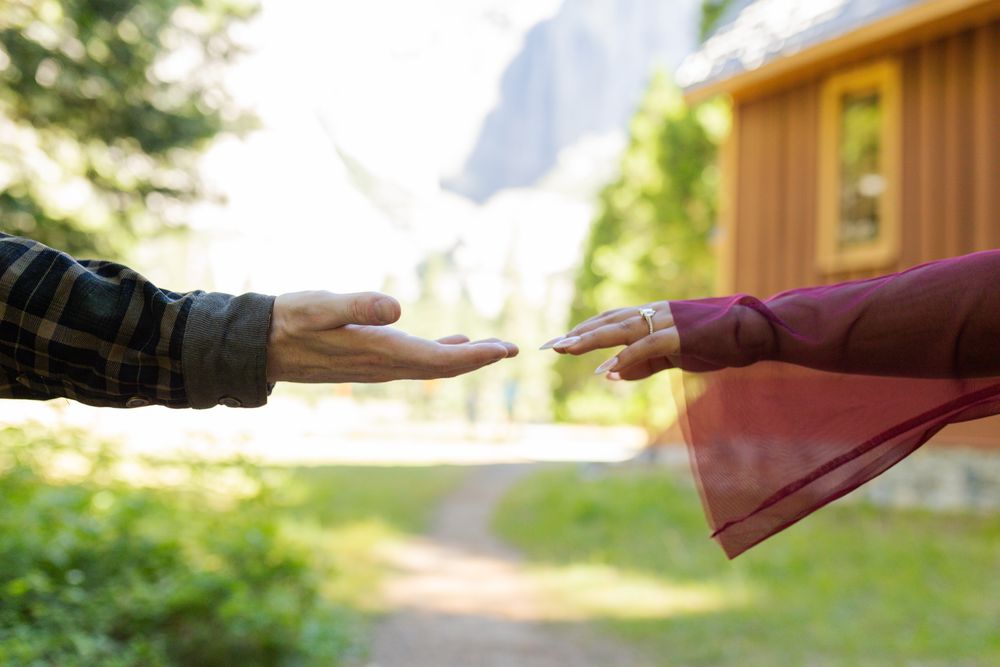 Two people reaching toward each other outdoors near a wooden cabin surrounded by lush greenery and mountains.