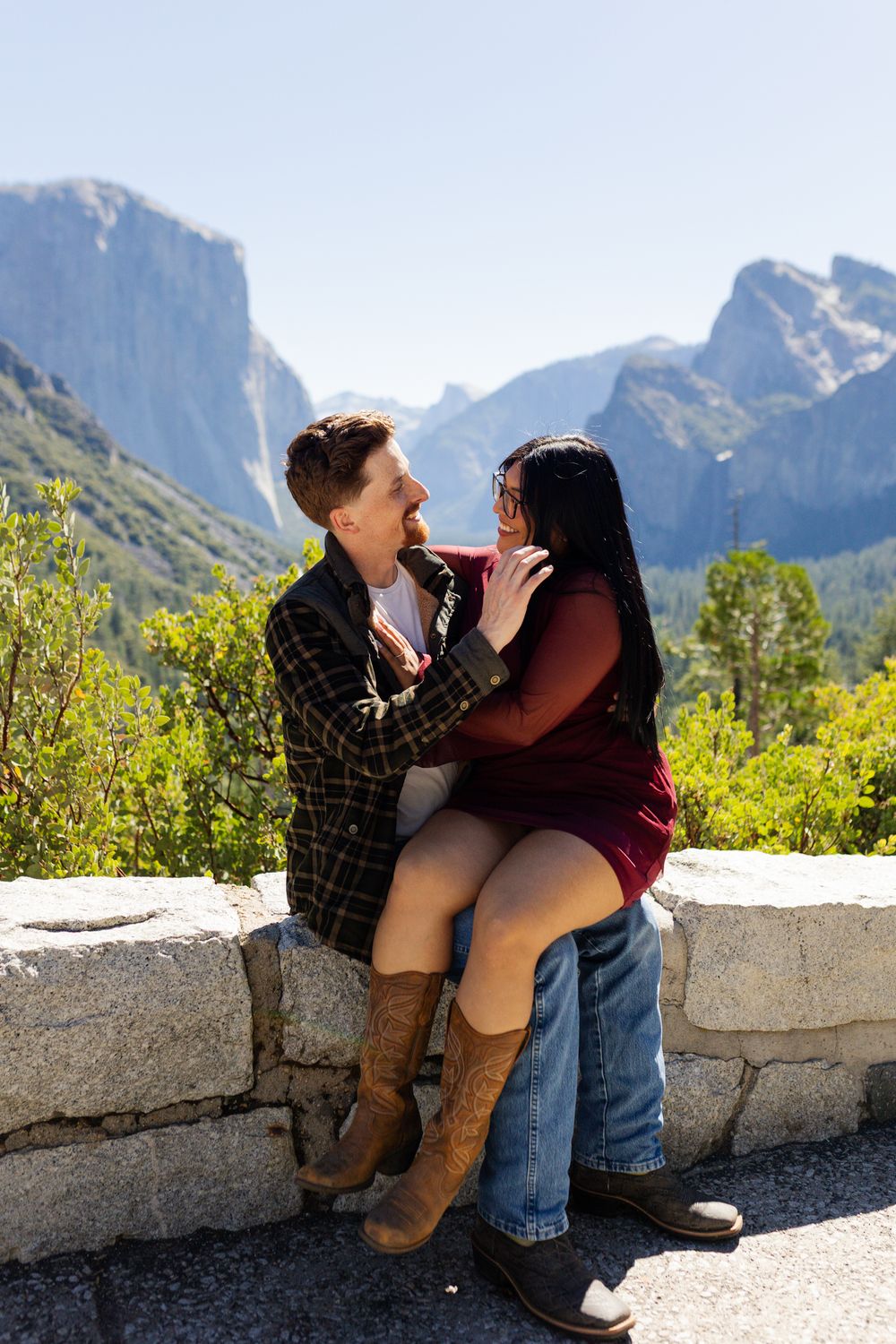 Couple sharing a romantic moment on a stone wall at Yosemite Valley with Half Dome in the background.