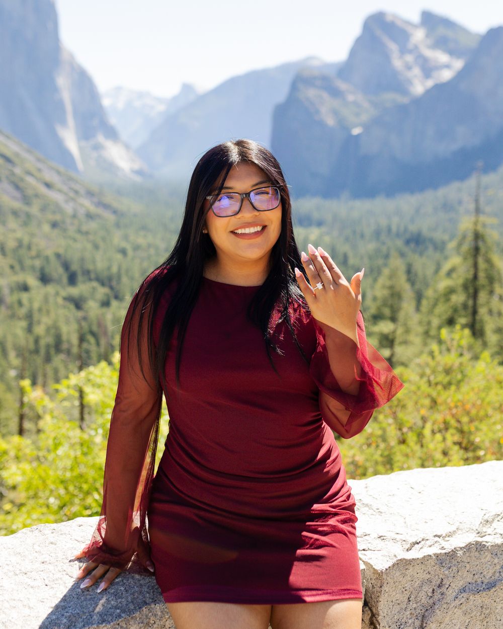 Woman in burgundy dress posing at Yosemite Valley overlook with mountains and forest backdrop.