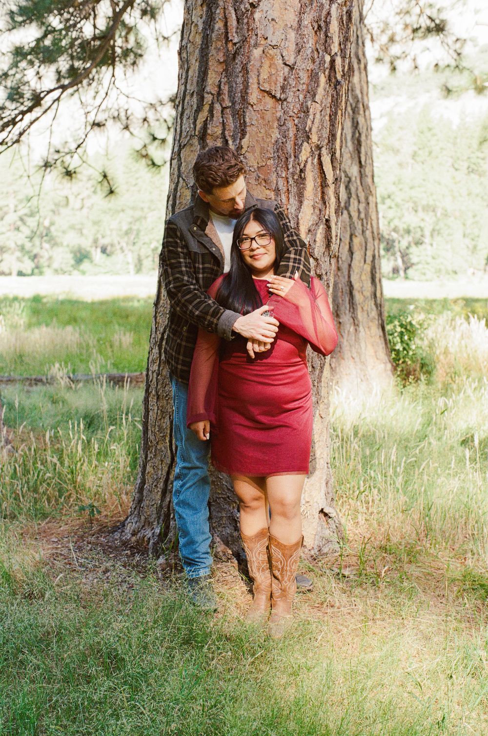 Couple embracing by a large tree in a park, woman in red dress and boots, man in plaid shirt and jeans.