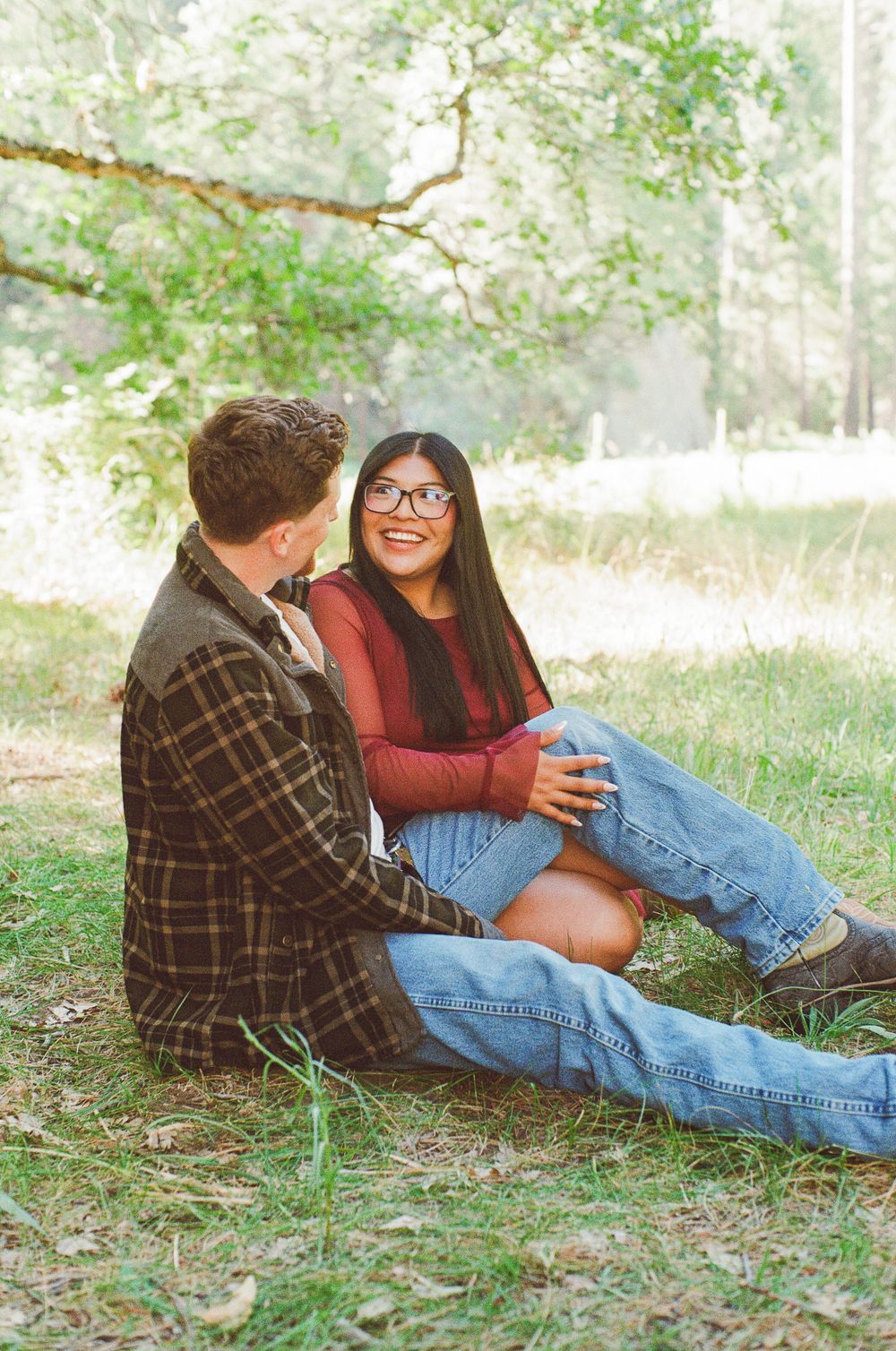 A smiling young woman in glasses and a red top sits with a man on grass beneath a tree in a sunlit park.