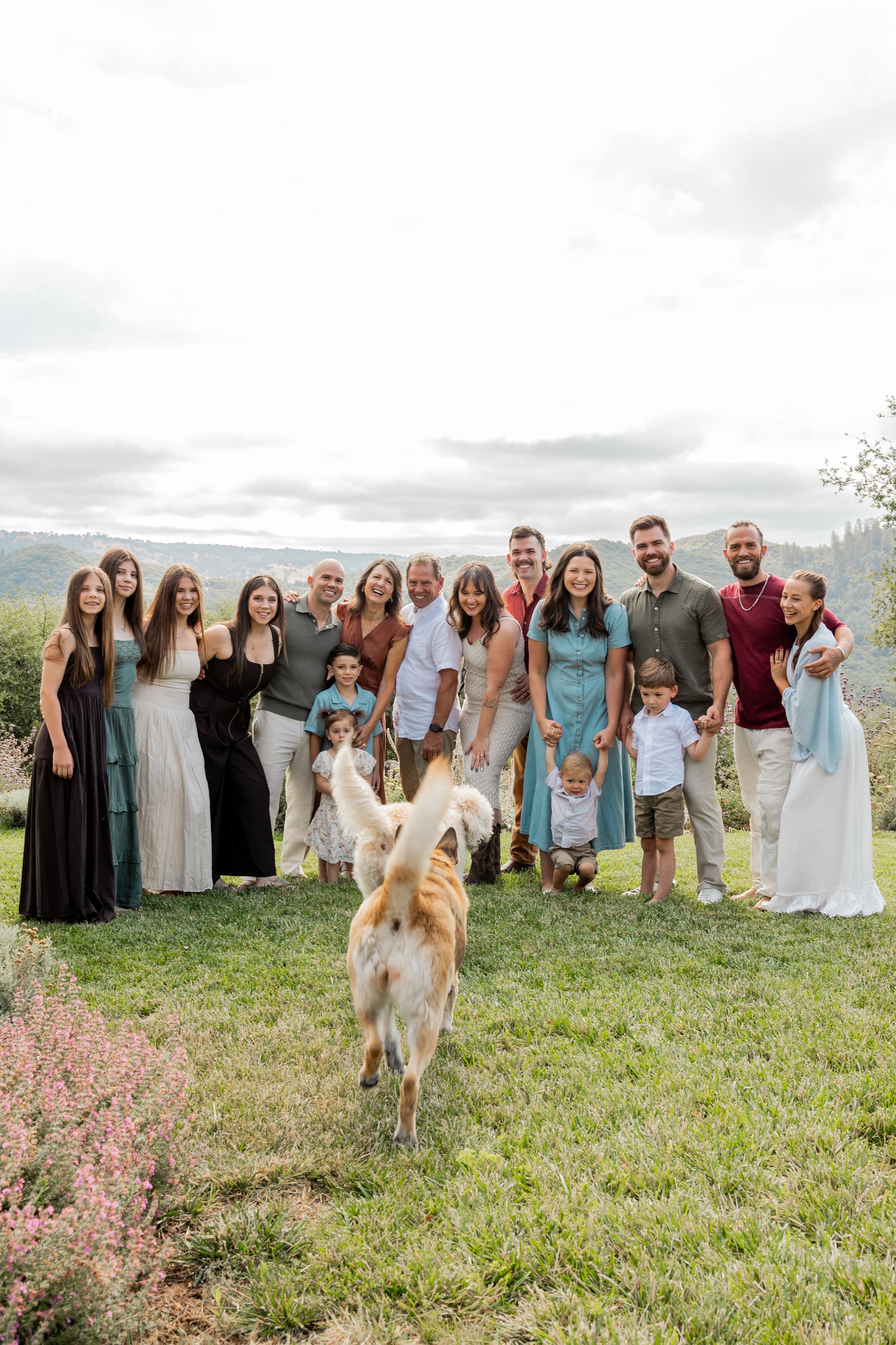 Large family group photo outdoors on green lawn with a dog running toward camera and scenic hills in background.