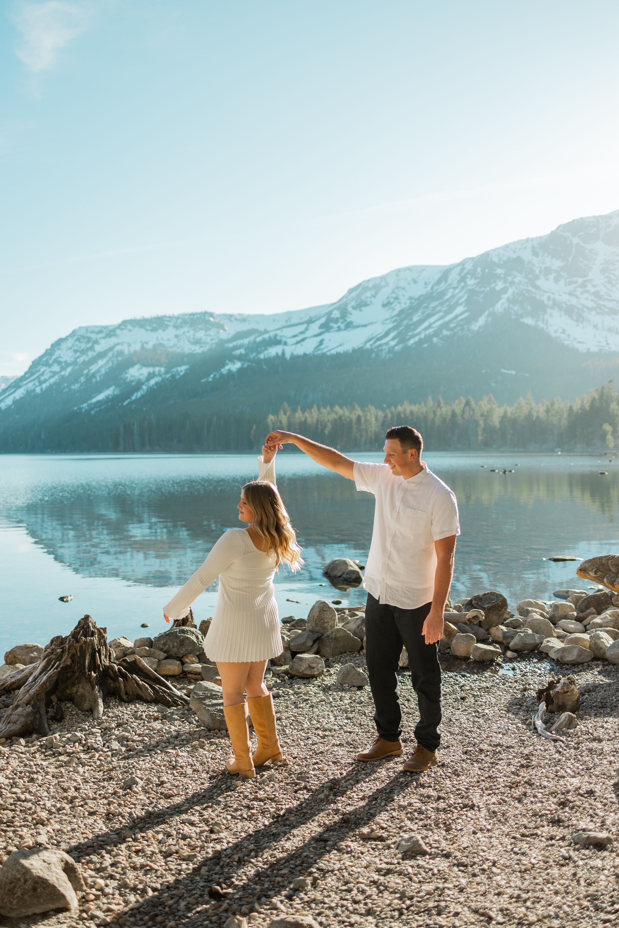 Couple dancing by a rocky mountain lake shore with snow-capped peaks and calm reflective water in background.