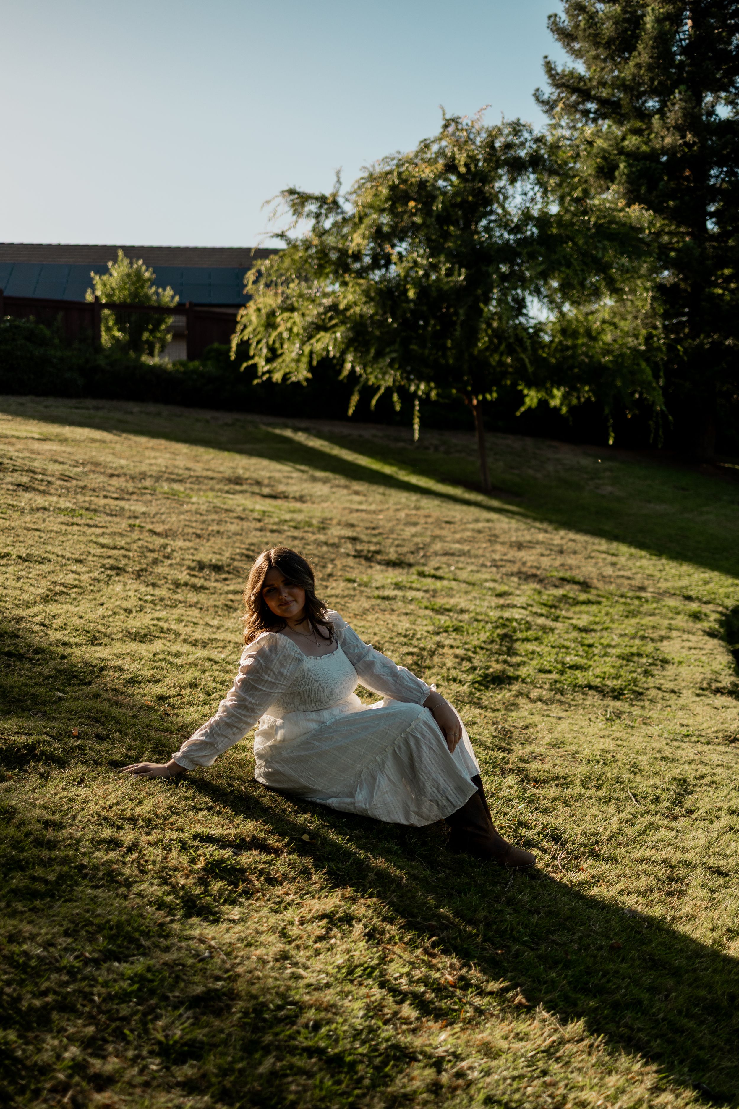 Woman in white lace dress sitting on sunlit grass in a garden with trees and a building in the background.