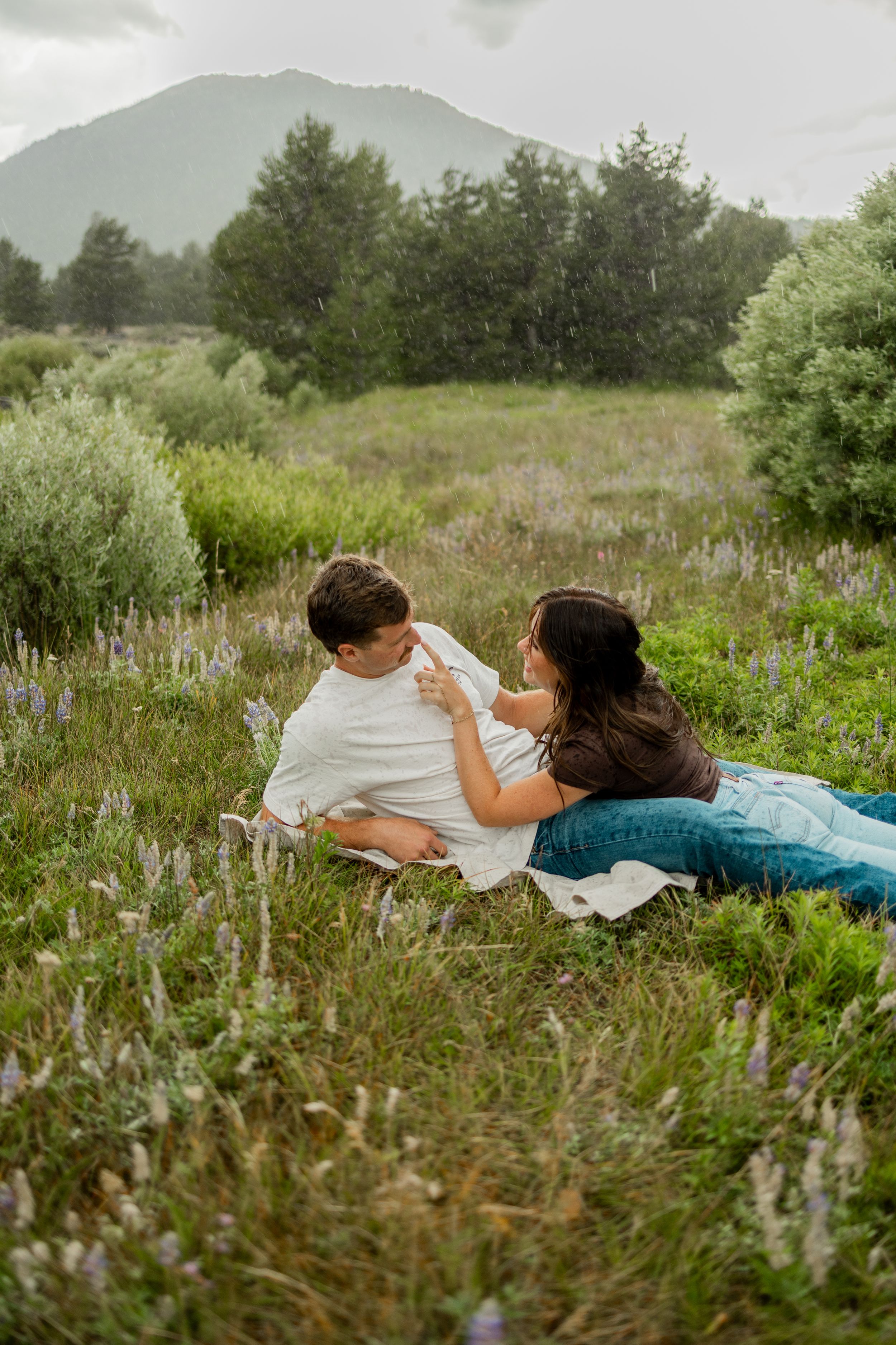 Couple relaxing together in a wildflower meadow with mountains and trees in the background on a rainy day.