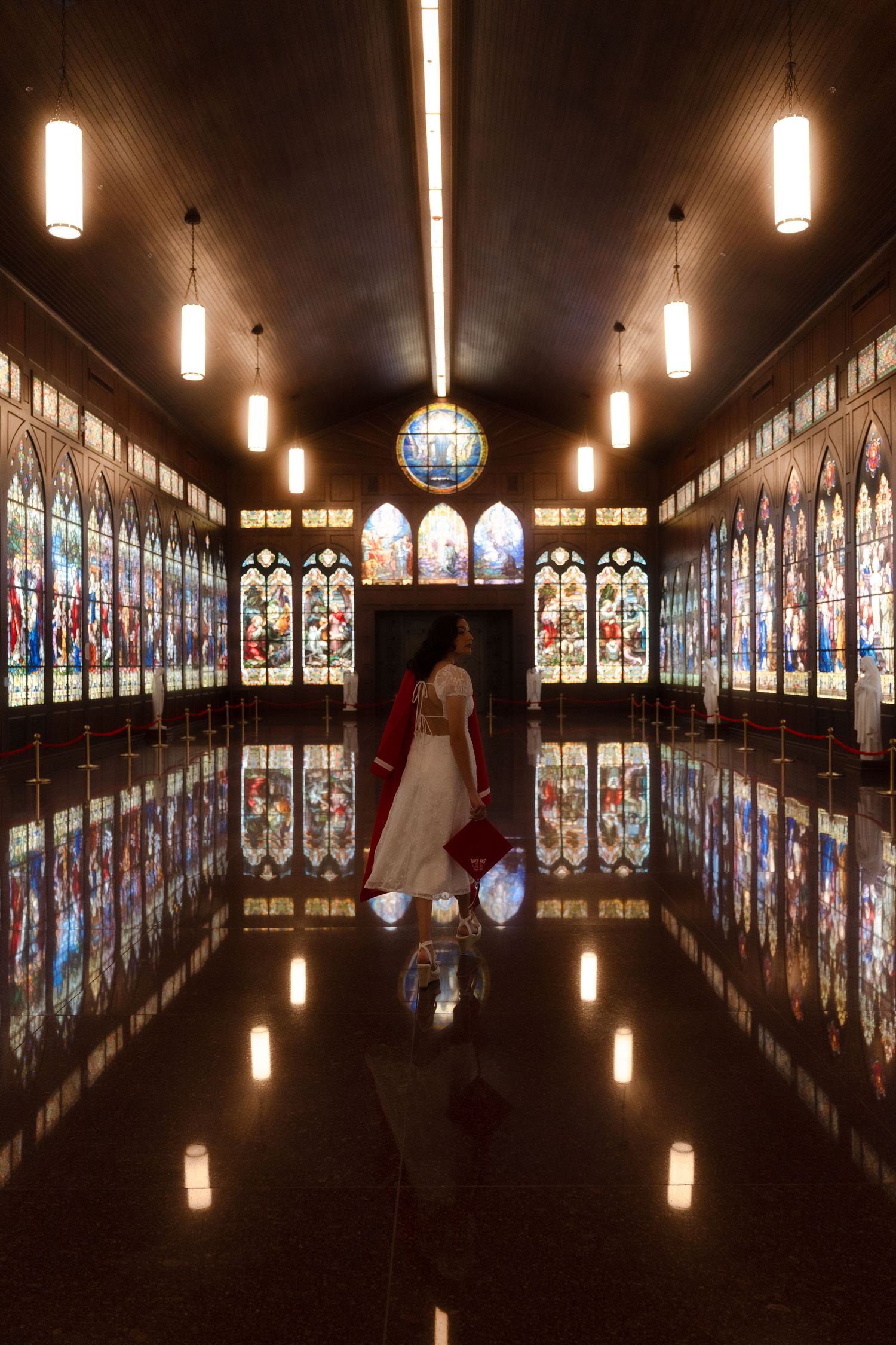 Bride in white gown with red cape walks through ornate chapel corridor lined with colorful stained glass windows and reflective floors.