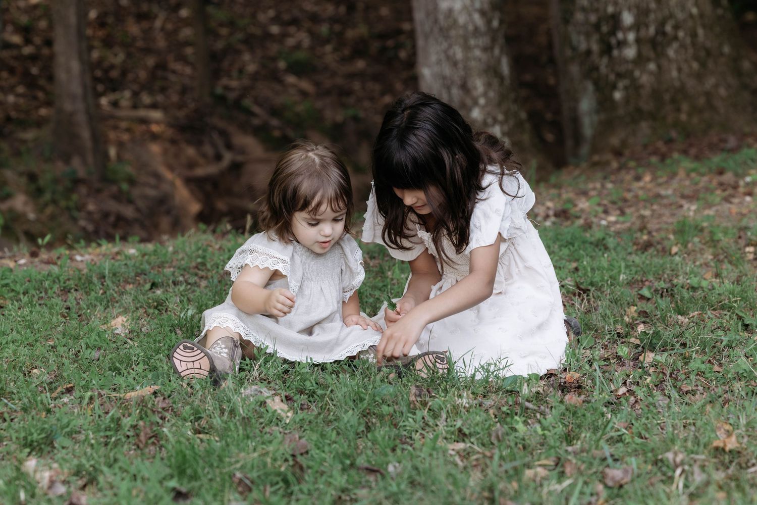 Pregnant woman in white dress walks with family holding hands on green grass with trees in background.