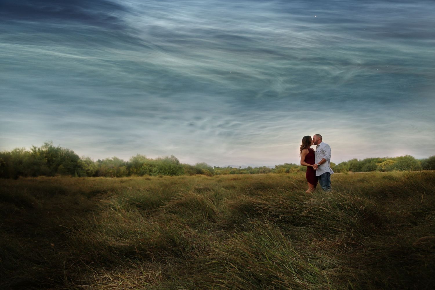 Couple shares a kiss in a grassy field beneath a dramatic blue sky with swirling wispy clouds.