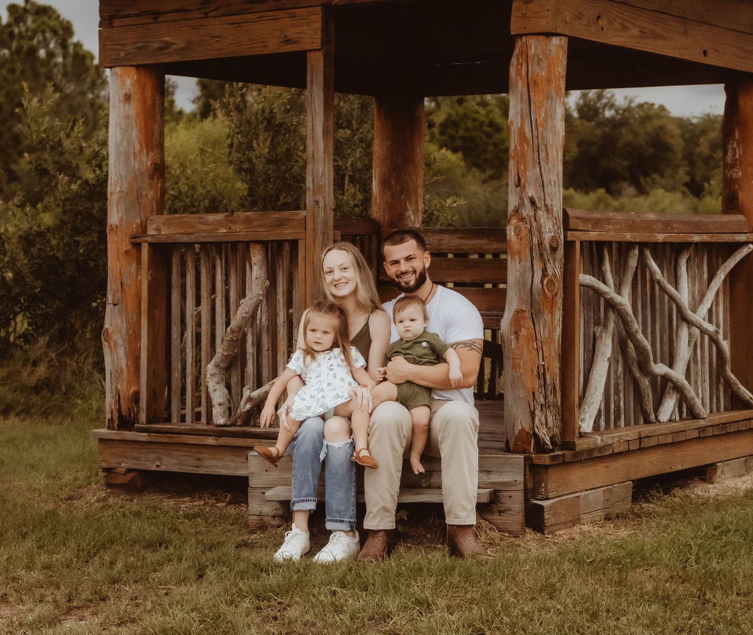 Family of four posing on rustic wooden gazebo steps in a green outdoor setting.