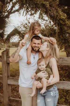 Happy family of four posing outdoors near a wooden fence, parents smiling with two young children in a park setting.