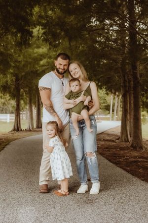 Happy family of four posing on a tree-lined path, parents holding young children outdoors.