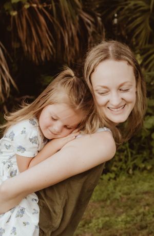 A smiling woman gives a young girl a piggyback ride outdoors, surrounded by lush green tropical foliage.