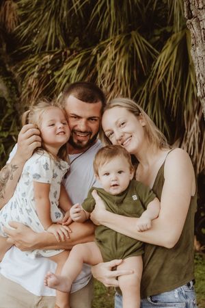 Happy young family with two toddlers smiling outdoors among lush tropical palm trees in a warm, candid portrait.