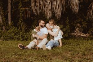 A father sits on grass with two young children in his lap, surrounded by lush tropical greenery.