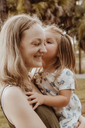 A young girl kisses her smiling mother on the cheek outdoors in a park setting.