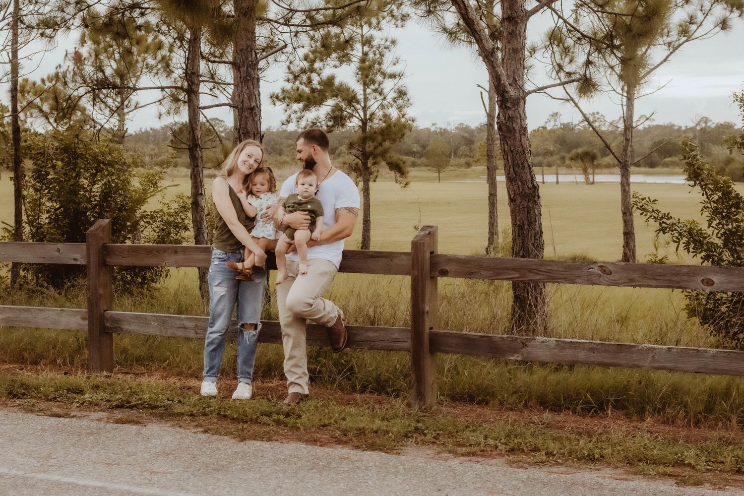 Family of three posing by rustic wooden fence surrounded by pine trees and green fields in a warm outdoor setting.