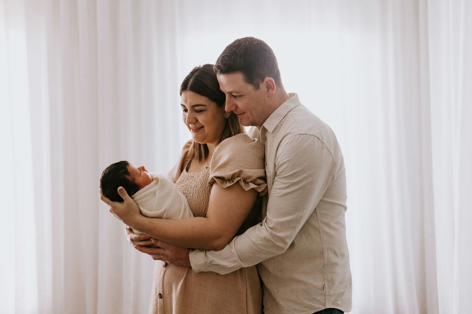 New parents lovingly gaze at their swaddled newborn baby in a bright, airy room with white curtains.