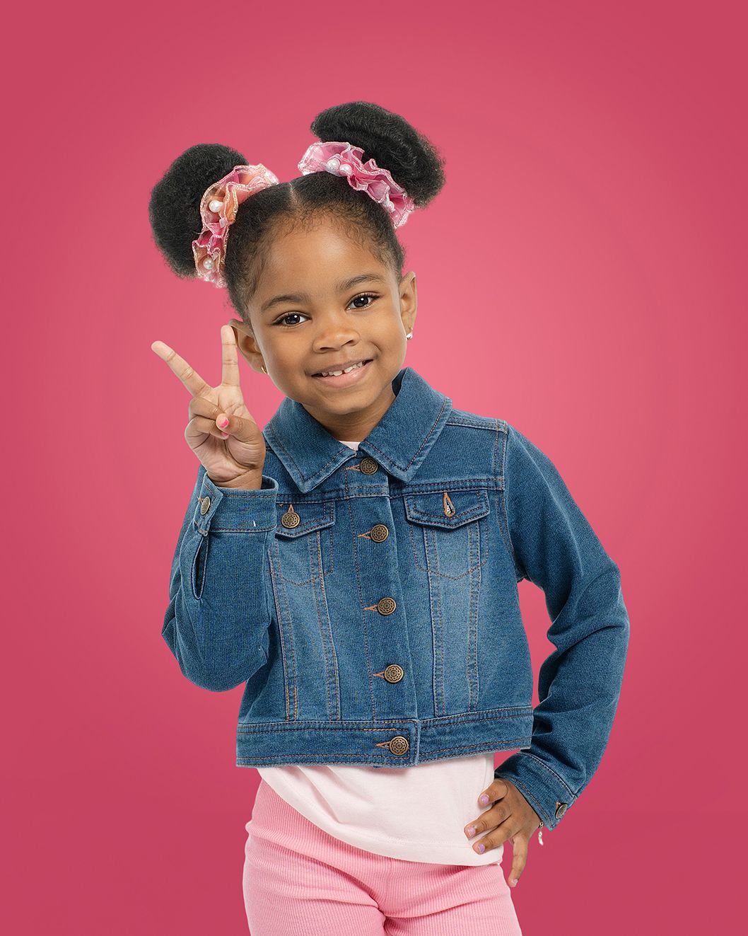 A young girl in a denim jacket and pink pants makes a peace sign against a pink background.