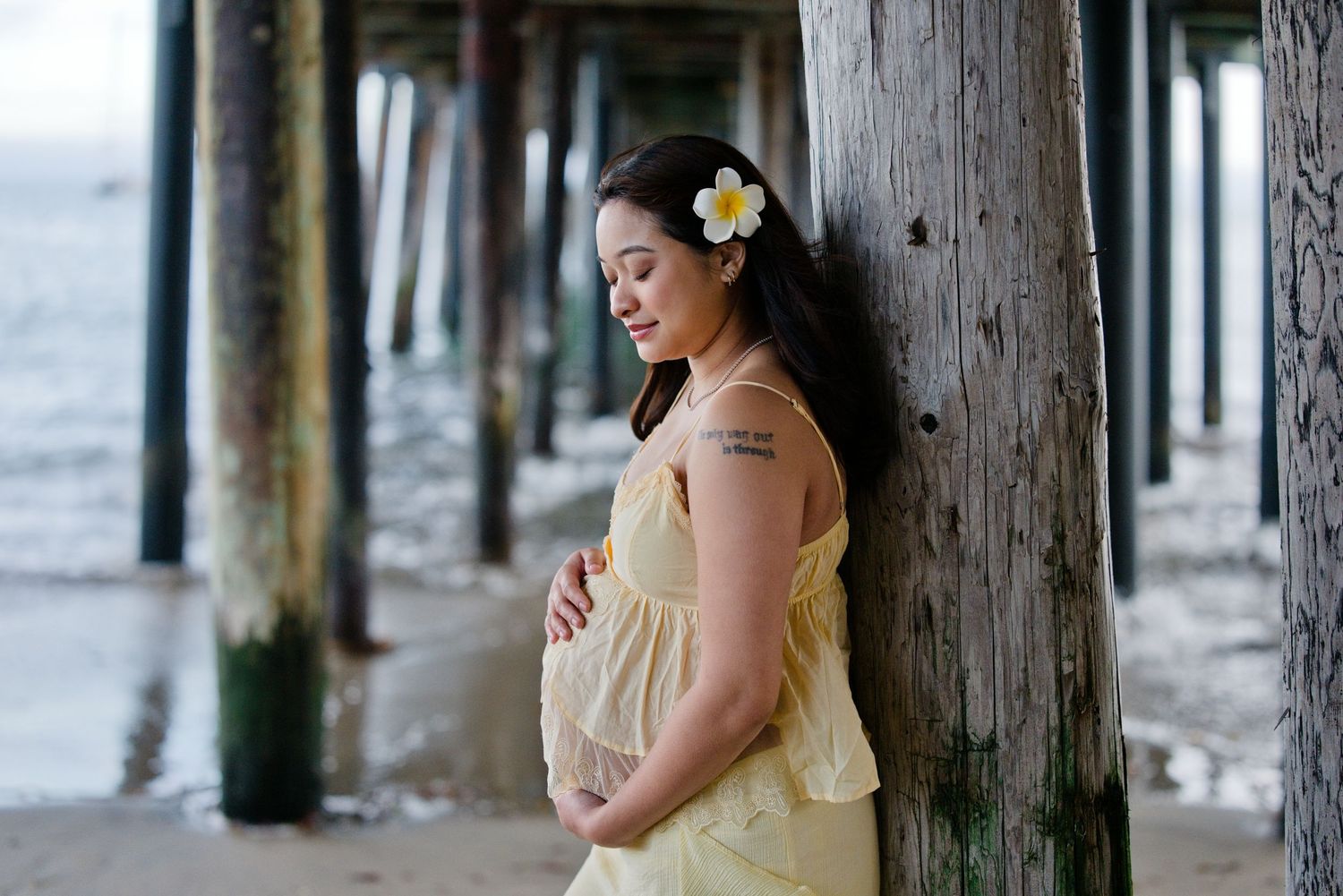 Pregnant woman in yellow dress holding belly under beach pier with flower in hair.