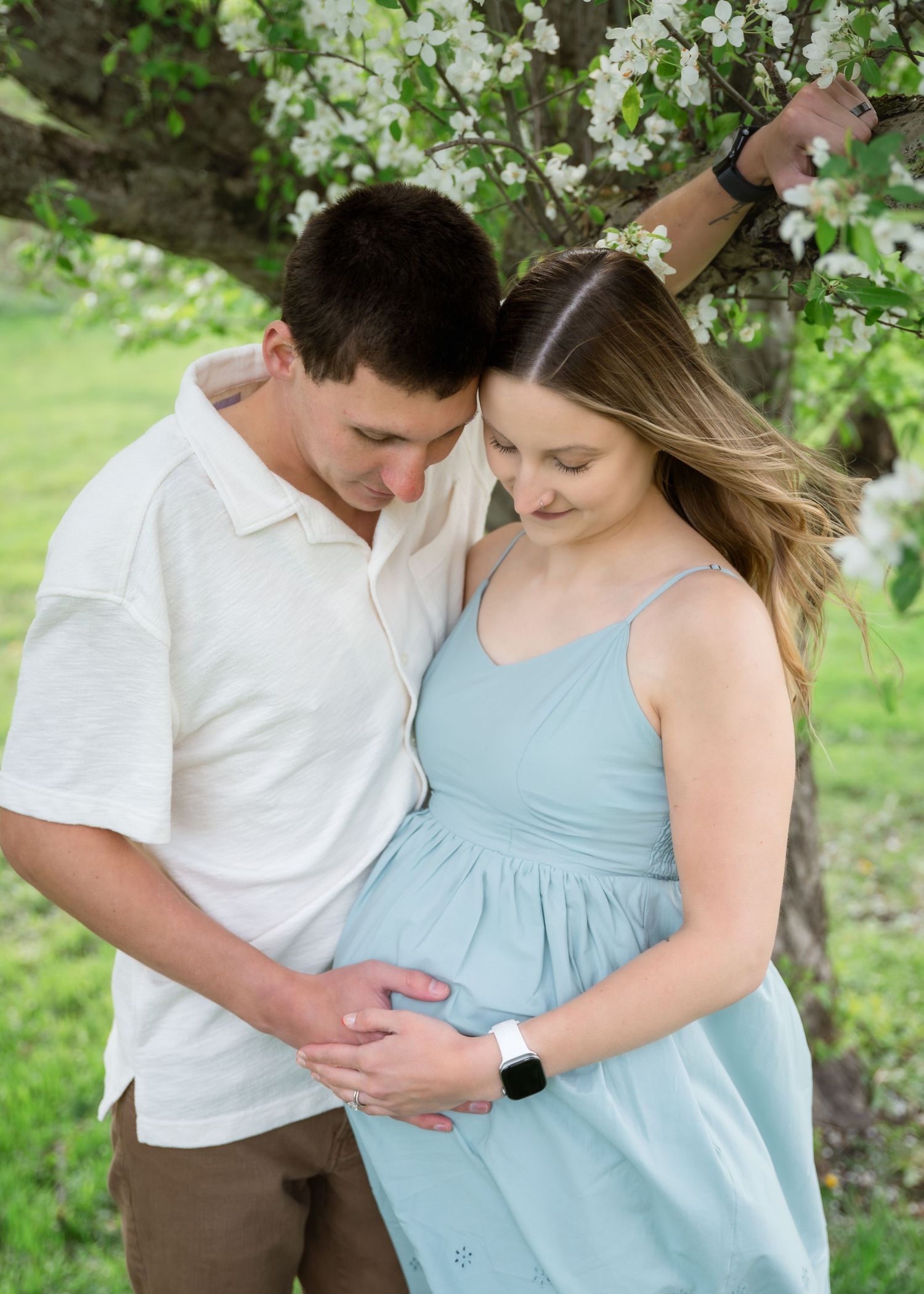 A couple tenderly cradles a pregnant belly under a blooming spring tree, the woman in a light blue maternity dress.