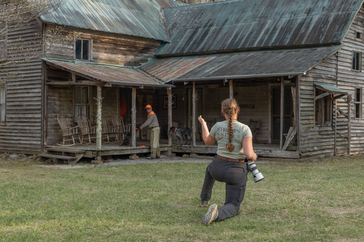 Photographer kneeling on grass capturing an old rustic wooden farmhouse with a person standing on the porch.