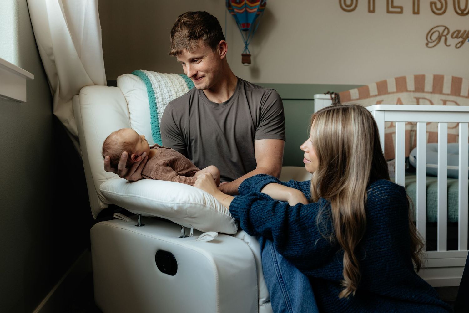 A couple sits on a white armchair holding their newborn baby, viewed through a doorway in a cozy home.