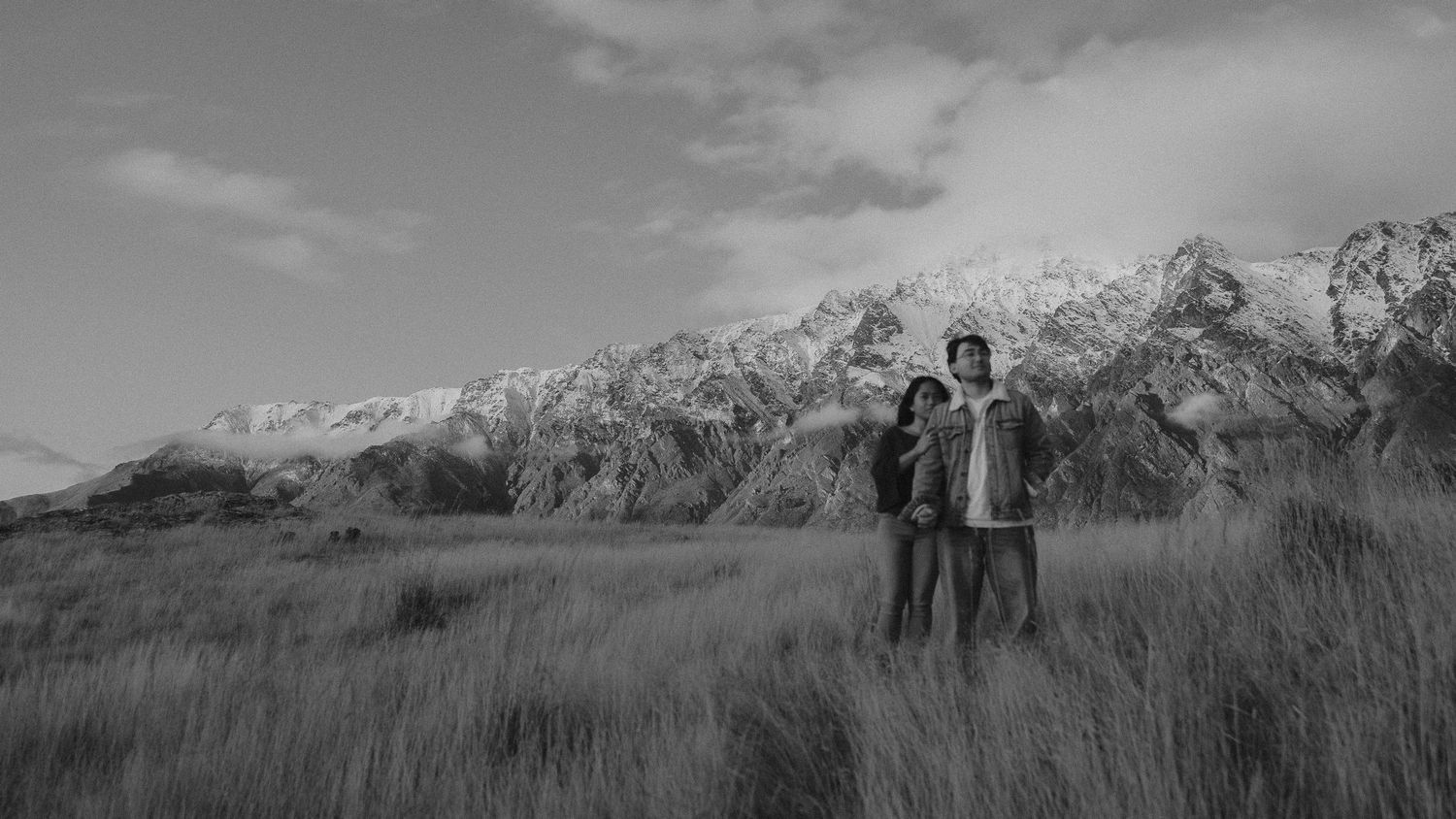 Two people standing in a grassy field with snow-capped mountains in the background, black and white photo.