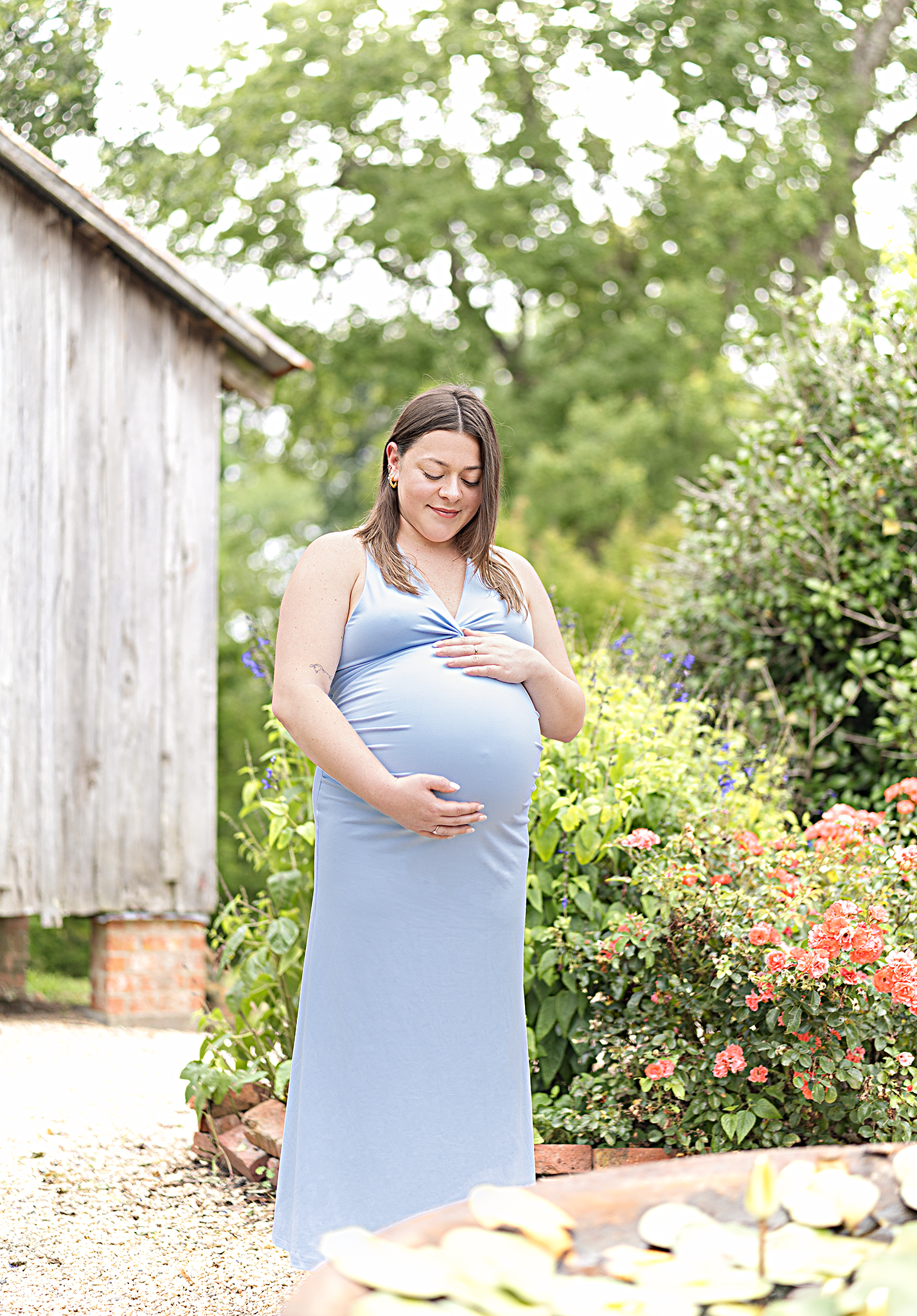 Pregnant woman in light blue maxi dress cradling baby bump in lush garden near rustic barn.