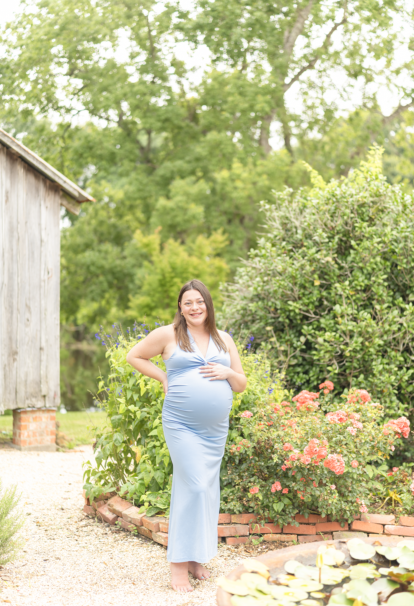 Happy pregnant woman in blue maxi dress posing barefoot in garden with colorful flowers and barn.