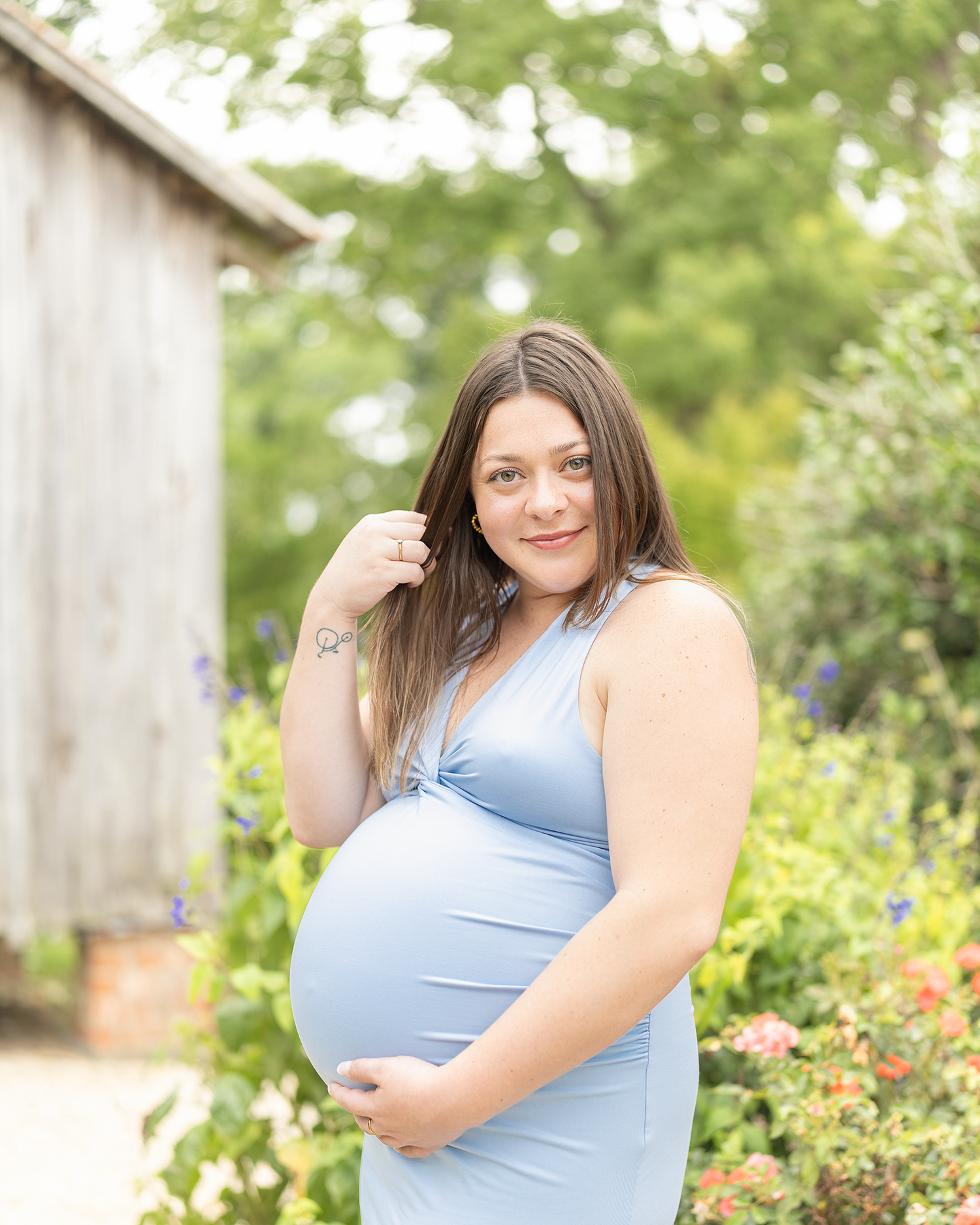 Pregnant woman in blue dress smiling and twirling hair while cradling bump near garden flowers.