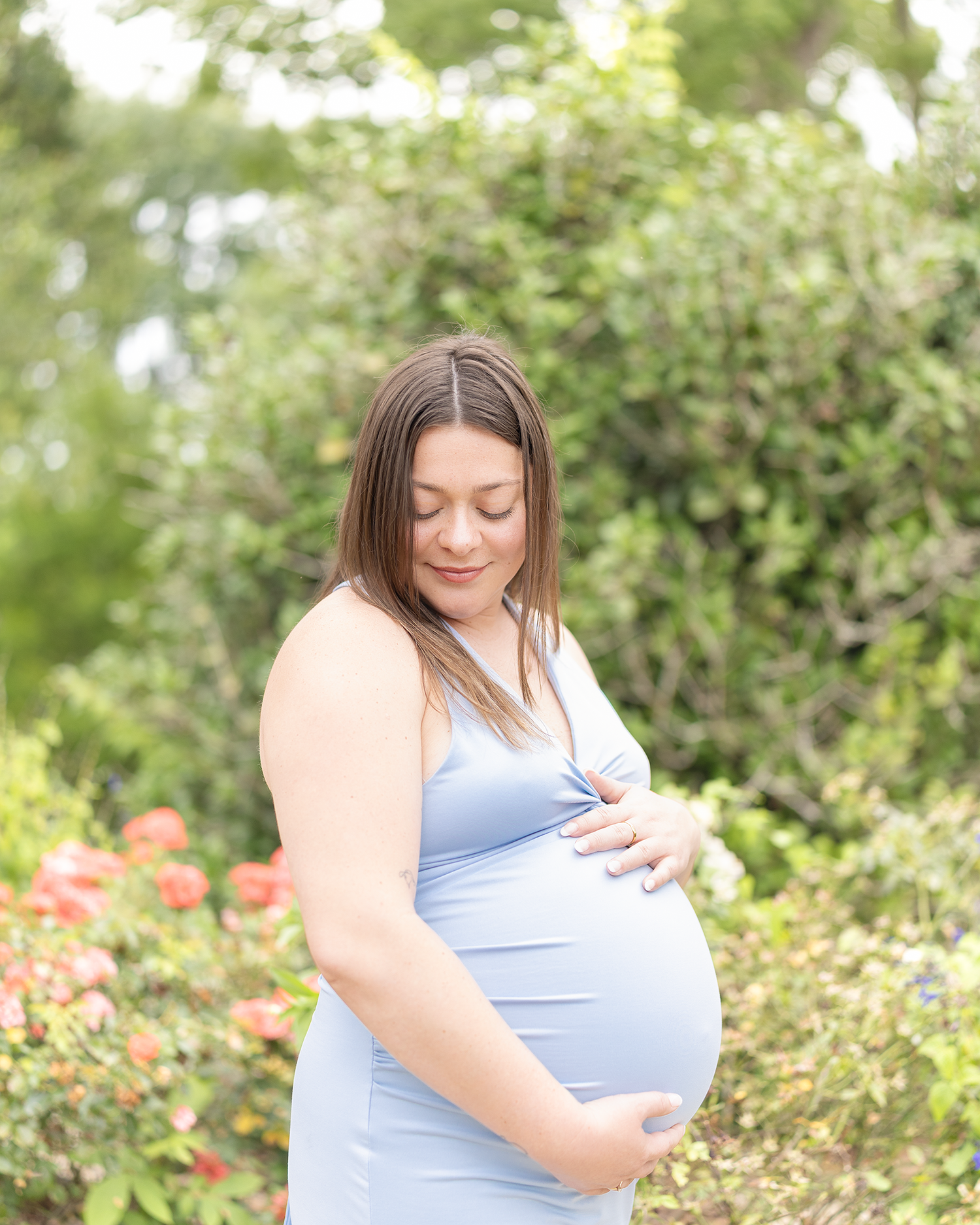 Pregnant woman in light blue dress looking down at baby bump surrounded by lush garden greenery.