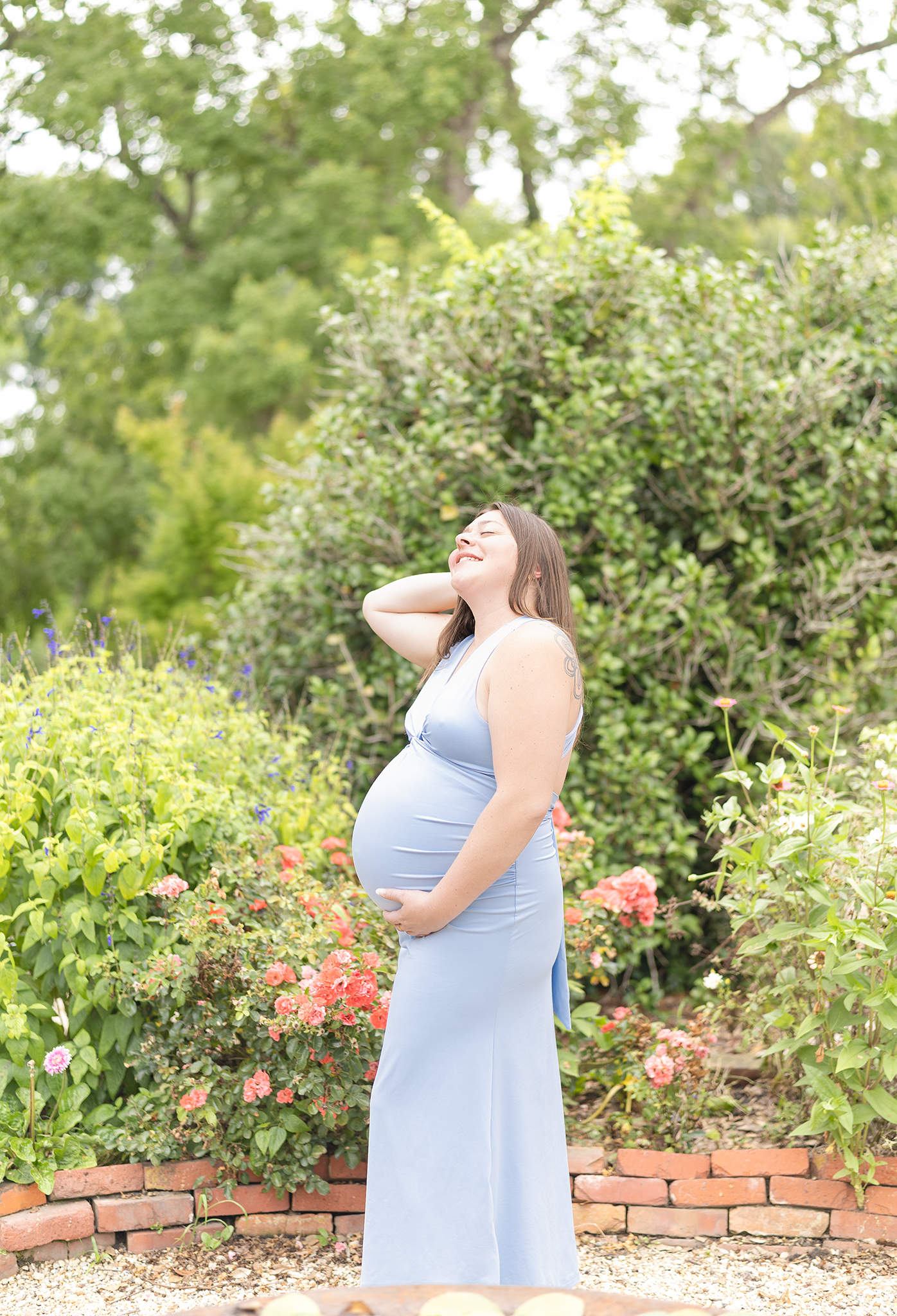 Joyful pregnant woman in blue maxi dress laughing with head tilted back in blooming garden.