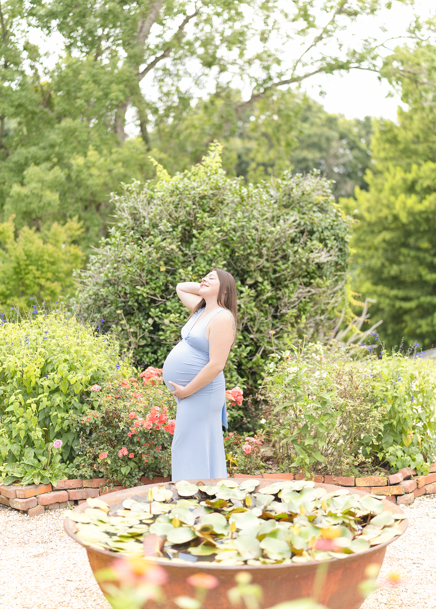 Pregnant woman in light blue dress standing beside lily pond fountain in lush garden setting.