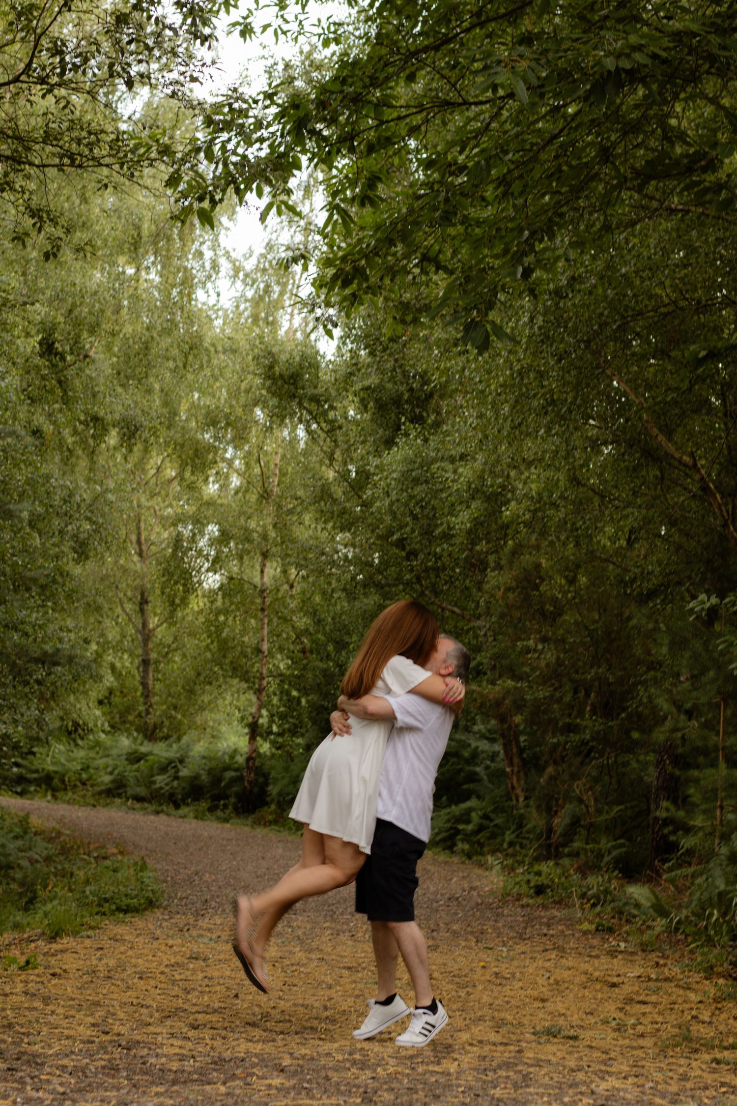 Couple embracing and spinning on a forest path surrounded by lush green trees in a romantic outdoor setting.