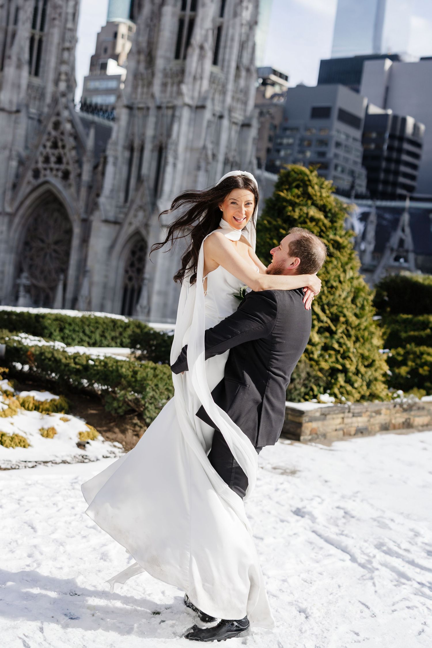 Groom lifts bride in black and white wedding gown outside snowy St. Patrick's Cathedral NYC.