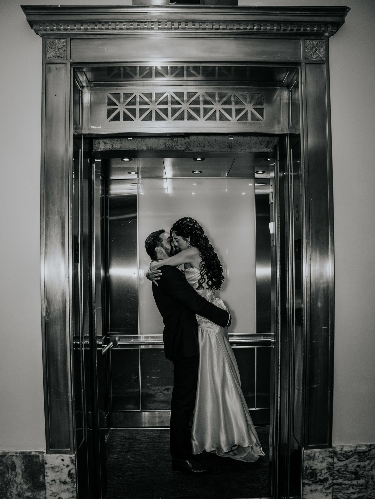 Black and white photo of a bride and groom sharing a kiss in an elegant elevator doorway on their wedding day.